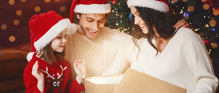 A young family wearing Santa hats and looking inside a glowing gift box.